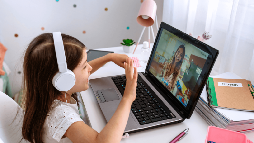 A little girl wearing a white headset waving to a woman on a video call.