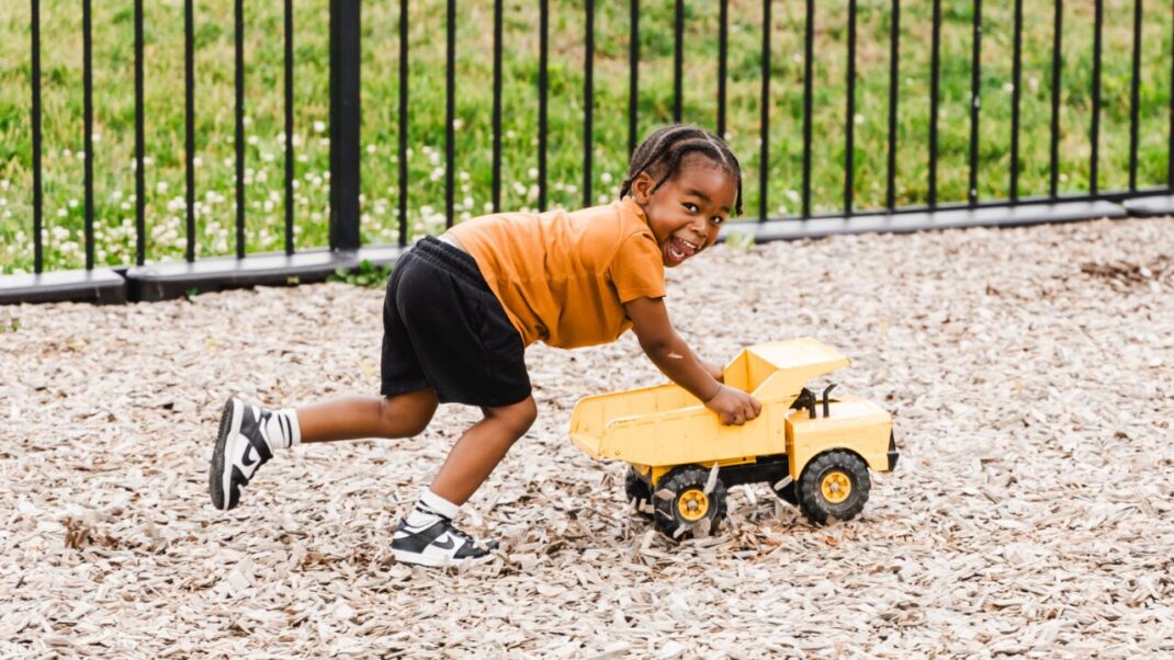 Child in an orange shirt pushing a toy truck outside.