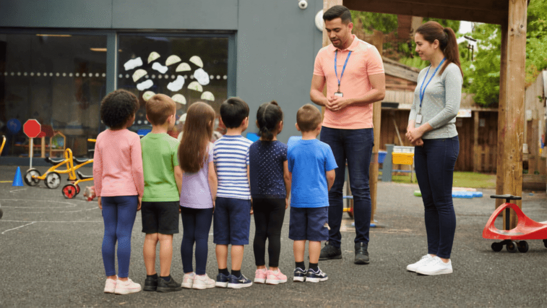Two adults stand facing a group of young children lined up outside near playground equipment at a daycare or school.